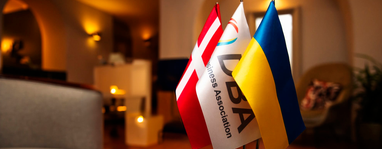 На зображенні на передньому плані розташовані три настільні прапорці: прапор Данії, прапор із логотипом «UDBA Business Association» та прапор України.