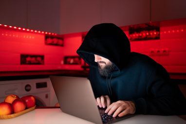 Хакер в черной толстовке с капюшоном на голове сидит за ноутбуком и печатает на клавиатуре