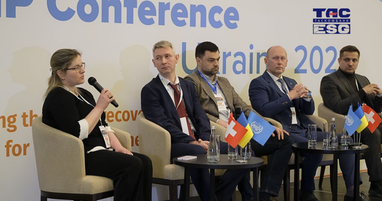 Спікерів (чотири чоловіки та жінка) сидять у кріслах на фоні банера заходу. Жінка ліворуч виступає з мікрофоном; на столиках перед учасниками стоять прапорці