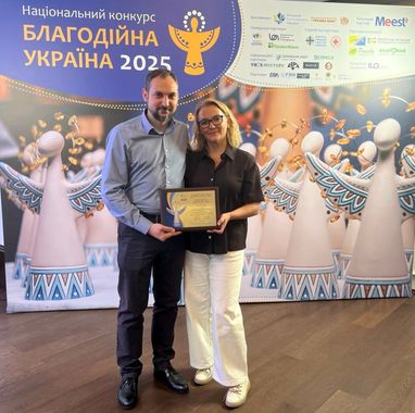 На фото изображены два человека, которые стоят на фоне крупного официального баннера и держат в руках диплом Национального конкурса «Благотворительная Украина-2025».
