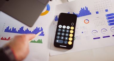 Cмартфон с открытым калькулятором, лежащий на финансовых документах с графиками и диаграммами, а рядом человек указывает на данные.