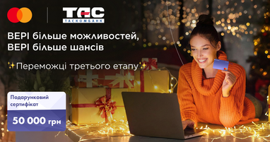 Усміхнена дівчина в помаранчевому светрі тримає платіжну картку біля ноутбука в оточенні подарунків та різдвяних вогників