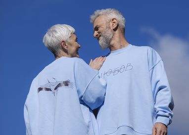 На фото пара похилого віку у блакитних світшотах з написом «Любов» та принтом журавлів на фоні синього неба. Чоловік та жінка дивляться один на одного з посмішкою.