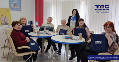 Клиентов пенсионного возраста сидят за круглыми столами в Сумском отделении № 51 Таскомбанка. Они держат в руках синие фирменные сумки с логотипом банка. За ними стоит улыбающаяся работница банка